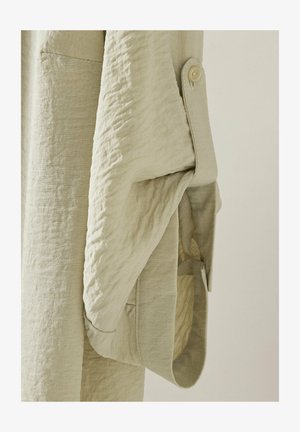 Camicia in lino color beige chiaro caratterizzata da un tessuto testurizzato, un bottone sul polsino e una tasca laterale con un drappeggio morbido e fluido.