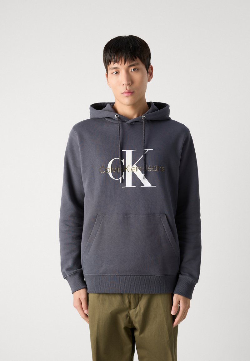 Sudadera de algodón gris con un bolsillo canguro en la parte frontal, capucha con cordón y un gran logo "CK" en blanco y dorado. Diseño sencillo, ajuste relajado.