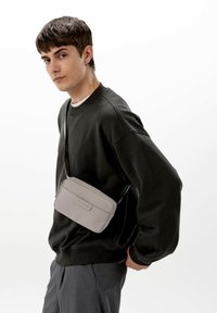 Šedá obdélníková kabelka přes rameno s hladkou texturou, zipovým uzávěrem a předním štítkem, nošena přes tmavý oversized svetr.