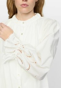 Weißes Blusenshirt mit hochgeschlossener Rüsche und Knopfleiste, das mit filigranen Spitzenverzierungen an den voluminösen Ärmeln versehen ist.