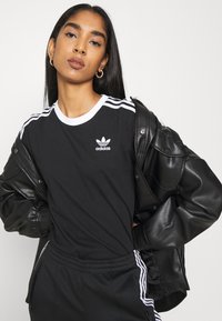 T-shirt de algodão preto com colarinho branco e três riscas brancas nos ombros. Logótipo da Adidas exibido no peito. Usado debaixo de uma jaqueta de couro preta.