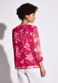 Blusa trasparente rosa con motivo floreale; maniche a tre quarti; schiena arricciata; indossata con pantaloni bianchi; texture liscia.