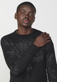Maglione nero in maglia con scollatura e polsini a coste, caratterizzato da motivi floreali argentati sul davanti. Texture morbida, vestibilità standard.