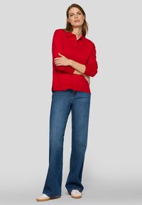 Rotes Langarm-Polo mit Kragen, bestehend aus drei Knöpfen am Ausschnitt. Kombiniert mit weit ausgestellten blauen Jeans und weißen Turnschuhen.