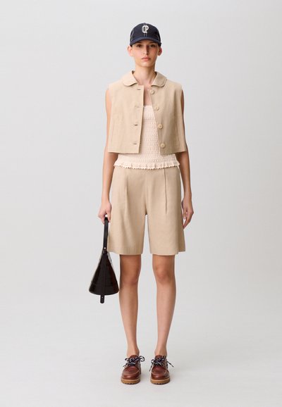 Donna che indossa un gilet beige senza maniche con bottoni, pantaloncini abbinati, top bianco testurizzato, cappellino blu navy, scarpe marroni e tiene in mano una borsa nera.