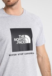 Camiseta de algodón gris con un logo cuadrado negro que lleva el texto en blanco "THE NORTH FACE" y "NEVER STOP EXPLORIN". Manga corta.