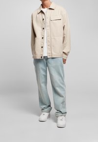 Mężczyzna ubrany w beżową marynarkę zapinaną na guziki, białą koszulkę, jasnoniebieskie dżinsy i białe sneakersy, stojący na szarym tle.