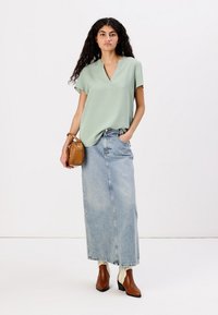 Blusa corta verde chiaro con maniche a cavallo e scollatura a V, abbinata a una lunga gonna di jeans sbiadita e stivaletti con tacco marroni, mentre regge una borsa piccola.
