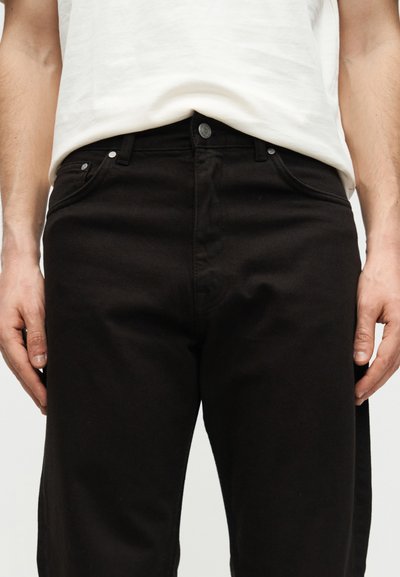 Άνδρας με μαύρο τζιν και λευκό πουκάμισο, με τα χέρια χαλαρά δίπλα στο σώμα, εμφανίζεται από τη μέση έως το μέσο του μηρού σε απλό φόντο.