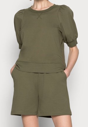 Kvinde iført olivengrøn kortærmet bluse med pufskuldre og matchende løse shorts med hænderne i lommerne mod en ensfarvet baggrund.