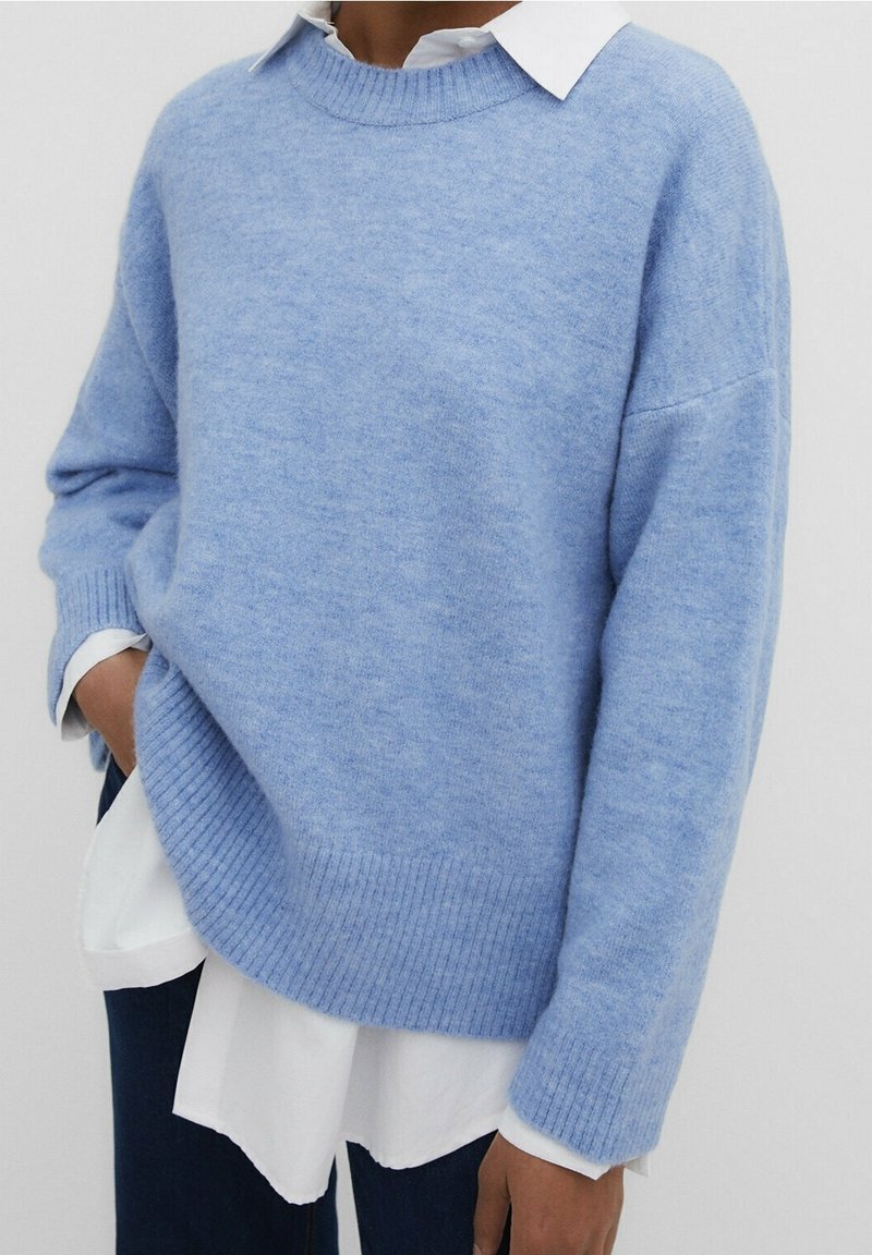Personne portant un pull tricoté léger bleu clair duveteux par-dessus une chemise blanche boutonnée avec un pantalon foncé, une main dans la poche, debout devant un fond uni.