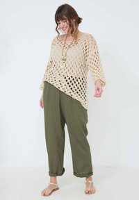 Pull en crochet beige au design en maille ajourée, associé à un pantalon vert ample. Le modèle porte des sandales et un collier de perles en couches.