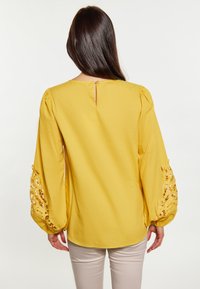 Blusa gialla con maniche a sbuffo, caratterizzata da dettagli di pizzo traforato sui polsini e da un piccolo forellino sulla schiena. Tessuto morbido.