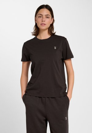 Jonge vrouw die een zwart T-shirt met korte mouwen en bijpassende broek met kleine chevronlogo's draagt, staand met handen in de zakken.