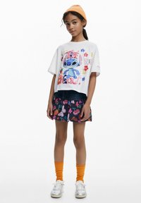 Vitt kort t-shirt med färggrafik av Stitch, parat med en svart kjol med livliga frukt- och blommönster. Orange stickad mössa, knästrumpor.