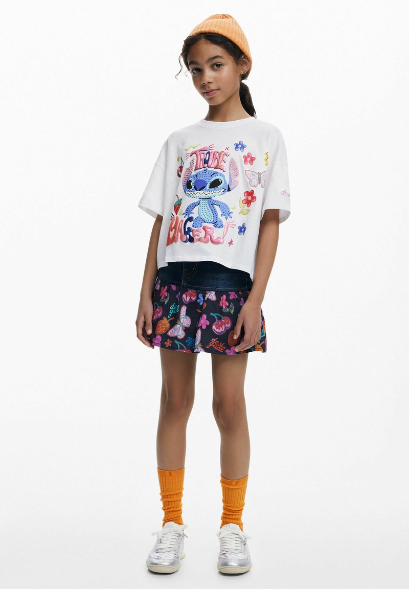 Vitt kort t-shirt med färggrafik av Stitch, parat med en svart kjol med livliga frukt- och blommönster. Orange stickad mössa, knästrumpor.