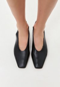 Ballerine in pelle nera con tacco basso, punta quadrata e lati aperti. Texture liscia con un design minimalista e senza hardware visibile.