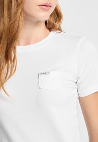 Baltas medvilnės marškinėliai su trumpomis rankovėmis, turintys kairėje krūtinės pusėje kišelį ir etiketę su užrašu "Guess Jeans." Lygus paviršius, paprastas dizainas.