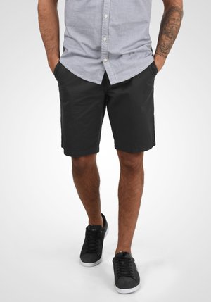 Black shorts made of cotton with side pockets, flat front, and a classic fit. Paired with black sneakers and a light blue button-up shirt.