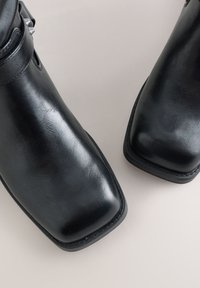 Schwarze Leder-Stiefeletten mit quadratischen Zehen und sichtbarer Naht auf hellem Hintergrund.