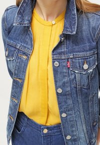 Giacca di jeans blu con dettagli in metallo e taschini sul petto, indossata sopra una camicia gialla a pieghe con collo rotondo.