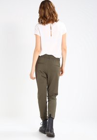 Witte korte mouwen top met opening aan de achterkant; olijfgroene tapered broek met elastische tailleband en twee achterzakken; zwarte enkellaarsjes.