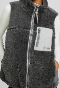 Szary polarowy kamizelka z wysokim kołnierzem, przednim zapięciem na zamek, teksturowanym materiałem oraz małą kieszenią na piersi z zamkiem błyskawicznym i detalem logo.