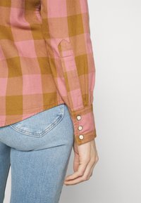 Personne portant un jean bleu et une chemise à manches longues à carreaux rose et marron avec trois boutons argentés sur le poignet.