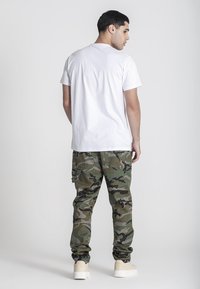 Camiseta blanca de algodón combinada con pantalones cargo de camuflaje verde, con bolsillos laterales y un corte relajado, sobre un fondo neutro.