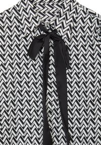 Blouse à motif zigzag noir et blanc avec un col et un nœud en satin noir noué au niveau de l'encolure.