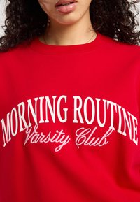 Rode sweatshirt met witte geborduurde tekst "MORNING ROUTINE Varsity Club." Het materiaal lijkt zacht en heeft een ronde halsontwerp.