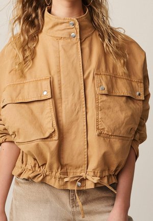 Femme portant une veste courte beige avec de grandes poches avant, des boutons-pression argentés, une ceinture à la taille et un pantalon marron clair texturé.