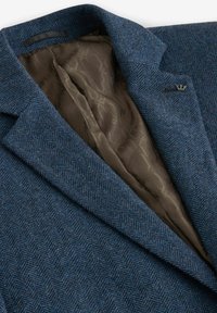 Blazer im blauen Fischgrätmuster mit einer strukturierten Außenfläche und einer glatten braunen Fütterung. Verfügt über Spitzrevers und subtile Hardwaredetails.