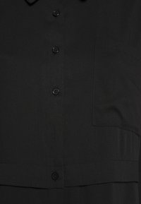 Camicia nera con colletto, dotata di tasca e chiusura con bottoni davanti. Realizzata in tessuto liscio con una sottile lucentezza e design strutturato.