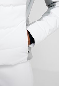 Vit puffjacka med grå detaljer, med en fickdetalj vid midjan, quiltad textur och en slimmad, modern design.