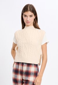 Cremfarbener ärmelloser Pullover mit Kabelstrickdesign, hohem Kragen und geripptem Saum, kombiniert mit karierter Hose in Rot- und Blautönen.
