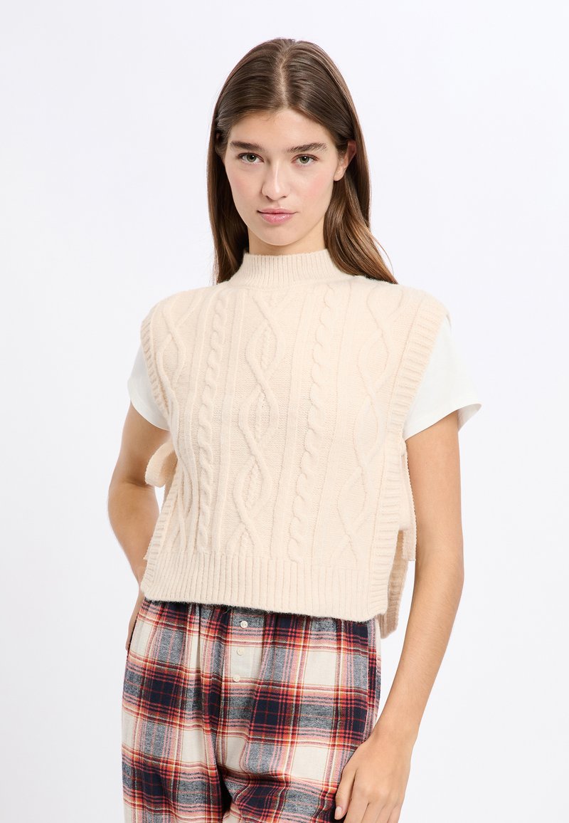Cremfarbener ärmelloser Pullover mit Kabelstrickdesign, hohem Kragen und geripptem Saum, kombiniert mit karierter Hose in Rot- und Blautönen.