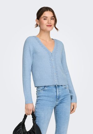 Femme portant un cardigan bleu clair boutonné et un jean, tenant un sac à main en cuir noir froncé avec une poignée courte.