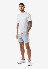 Man with tattoos stands wearing light gray short-sleeve shirt, matching shorts with white side panels, and white athletic shoes.