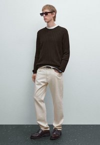 Junger Mann, der eine Schildpatt-Sonnenbrille, einen dunkel gerippten Pullover, helle Hosen und braune Lederschuhe trägt und mit einer Hand in der Tasche steht.