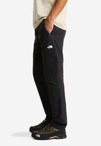 Pantalones negros de exterior hechos de material duradero. Cuentan con bolsillos con cremallera, un corte holgado y un logo en el muslo. Se llevan con zapatillas de senderismo.