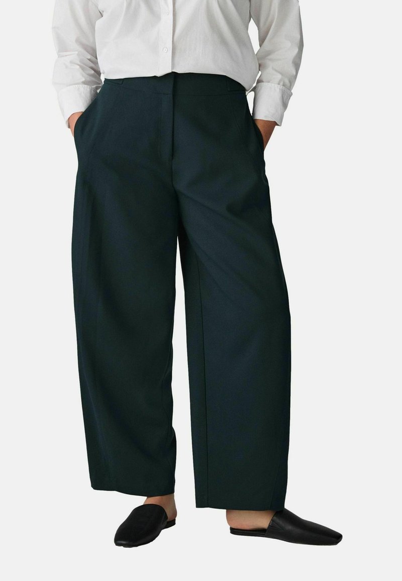 Weite, dunkelgrüne Hose mit hoher Taille, seitlichen Taschen und glatter Textur, kombiniert mit schwarzen Slippern.