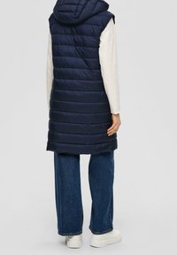 Lange marineblaue Steppweste mit Kapuze und gesteppter Struktur. Trägt man über einem hellfarbenen Langarmshirt, kombiniert mit locker sitzenden Jeans und weißen Schuhen.