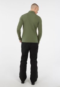 Uomo in piedi di spalle, che indossa una camicia a maniche lunghe verde oliva e pantaloni neri, su uno sfondo bianco semplice.