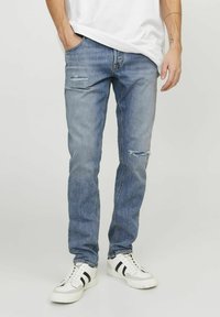 Jeans de denim azul claro con un corte holgado, que presentan parches desgastados, desvanecimiento y un diseño clásico de cinco bolsillos, llevados con zapatillas blancas.
