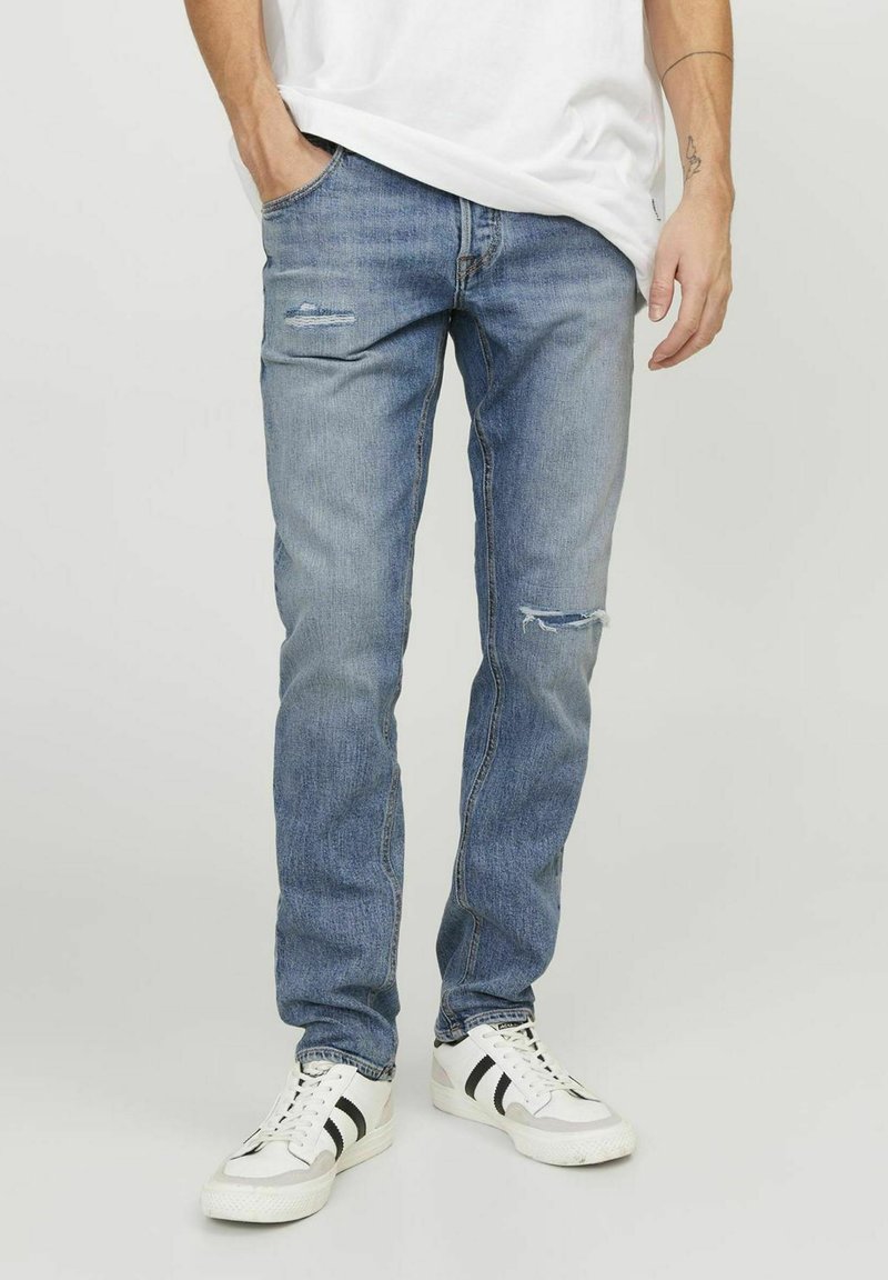 Jeans de denim azul claro con un corte holgado, que presentan parches desgastados, desvanecimiento y un diseño clásico de cinco bolsillos, llevados con zapatillas blancas.