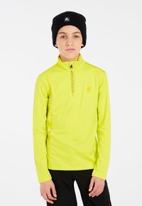 Camicia a maniche lunghe con colletto alto in tessuto giallo brillante, dotata di zip nera e logo. Indossata con un berretto nero. Sfondo semplice.