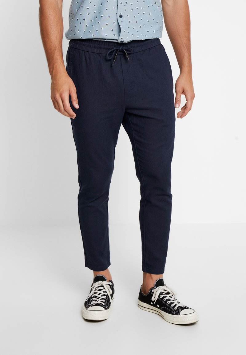 Man in donkerblauwe sweatpants met trekkoord, zwart-witte sneakers en een lichtblauwe korte mouw blouse met kleine navy patronen.