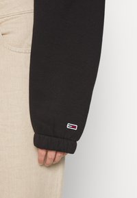 Sudadera negra de manga larga con puños elásticos, que presenta un pequeño logo bordado en la manga. Llevado sobre pantalones beige.