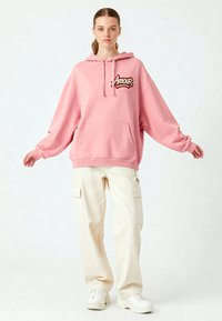 Felpa oversize rosa con tasca frontale e ricamo "Amour"; abbinata a pantaloni cargo color crema e sneakers bianche. Texture liscia.